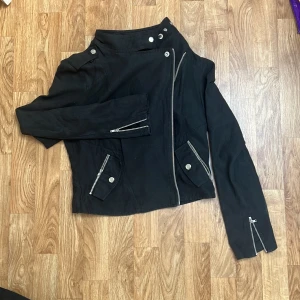 Svart tunn bikerjacka Saints & Mortals - Svart tunn bikerjacka från Saints & Mortals i storlek S. I mockaimitation. Jackan har coola silvriga dragkedjor, knappar och detaljer vid kragen. Asymmetrisk dragkedja framtill. Använd fåtal gånger.