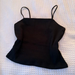 Svart ribbad linnetopp med smala band - Snygg svart linnetopp med smala axelband och ribbad struktur. Toppen har en enkel, croppad passform som ger en cool och stilren vibe. Perfekt att matcha med jeans eller kjol för en trendig look.