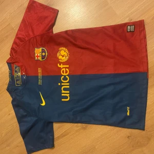 FC Barcelona Messi 10 Nike fotbollströja - Säljer en ikonisk FC Barcelona fotbollströja från Nike med Messi 10 på ryggen. Tröjan har klassiska röda och mörkblå paneler, gula detaljer och klubbmärke på bröstet. Materialet är lätt och andas, perfekt för match eller träning. UNICEF-logga och finaldetaljer från 2009.