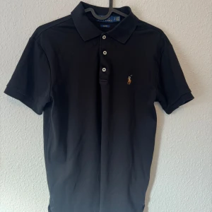 Svart pikétröja från Ralph Lauren - Snygg svart pikétröja från Ralph Lauren med klassisk krage och tre knappar framtill. Ikoniska broderade loggan på bröstet. Perfekt för dig som gillar stilren och enkel design. Tröjan är i mjuk bomull och har korta ärmar.