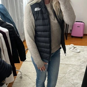 Svart dunväst från The North Face - Säljer en svart dunväst från The North Face med quiltad design och logga på bröstet. Västen har en klassisk, rak passform och dragkedja framtill. Perfekt att slänga över en hoodie eller stickad tröja för extra värme. Enkel och stilren look.