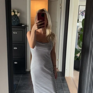 Randig vit och svart långklänning - Säljer en snygg långklänning med smala svarta ränder på vit botten. Klänningen är ärmlös och har en figurnära passform med rundad halsringning. Perfekt för dig som gillar enkel och stilren design med en modern touch.