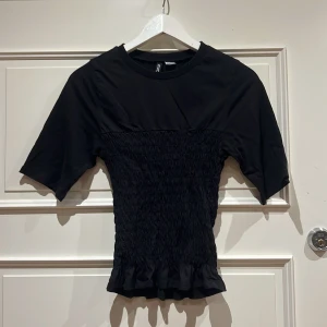 Svart smockad topp från H&M Divided XS - Svart topp från H&M Divided i storlek XS. Toppen har smockad midja och volangkant nertill, med korta ärmar och rund halsringning. Materialet är mjukt och stretchigt, perfekt för en trendig look.