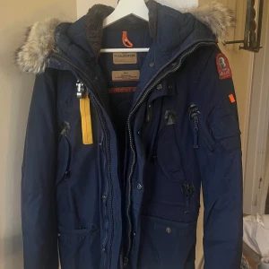 Blå Parajumpers parkajacka med päls - Säljer en mörkblå Parajumper med fluffig päls på huvan, coola detaljer som gul rem och flera fickor. Jackan har quiltad insida, dragkedja och knappar framtill. Perfekt för kalla vinterdagar och riktigt snygg stil. Mycket fint skick 