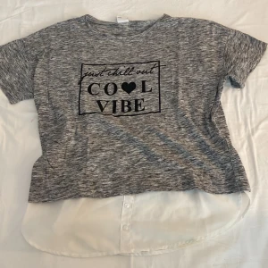 Grå t-shirt med text från Lindex - Snygg gråmelerad t-shirt från Lindex med svart tryck 'Just chill out COOL VIBE' och hjärta på bröstet. T-shirten har korta ärmar och en vit, skjortlik nederdel med knappar som ger en cool lager-på-lager look. Perfekt för dig som gillar streetstyle.