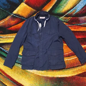 Marinblå vind feildjacket ifrån These Glory Days i väldigt bra skick  - Snygg marinblå vind feildjacket  ifrån These Glory Days i storlek medium. Jackan har hög krage med vit snörning, fyra fickor framtill med lock och knappar samt diskreta vita detaljer på kragen. Perfekt för dig som gillar stilren och funktionell design. I väldigt bra skick skick 9/10 pågrund av att man kan se att jackan är lite använd  DM för fler bilder eller frågor 🎬®️☔️