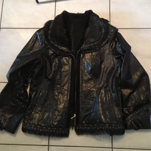 Svart skinnjacka med päls och pärlor - Snygg svart skinnjacka med bred krage i päls och pärldetaljer längs kragen och ärmsluten. Jackan har dragkedja framtill och raka fickor. Materialet är glansigt och har en cool struktur, perfekt för dig som gillar att sticka ut med detaljer.