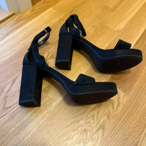 Svarta mockasandaler med klack från Pace - Snygga svarta sandaler från Pace i mjuk mocka med bred klack och plattform. Skorna har öppen tå, justerbart spänne runt ankeln och stilren design som ger en cool vibe. Perfekta för dig som gillar att sticka ut med chunky klackar och minimalistisk look.