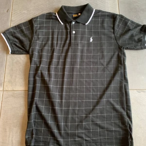 Polo Ralph lauren piké - Svart t-shirt från Polo Ralph Lauren med vit rutigt mönster och klassisk pikékrage. T-shirten har korta ärmar, vita detaljer på krage och ärmslut samt den ikoniska broderade loggan på bröstet. Perfekt för dig som gillar stilren och sportig look. Storlek L men passar M