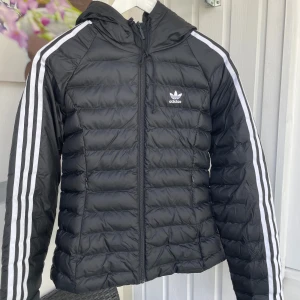 Svart dunjacka från Adidas med huva - Svart dunjacka från Adidas med klassiska vita ränder längs ärmarna och Adidas-logga på bröstet. Jackan har huva, dragkedja framtill och quiltad design. Perfekt för kyliga dagar och riktigt snygg streetstil.