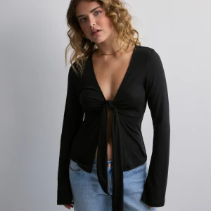 Svart omlottopp med knyt framtill - Svart långärmad omlottopp med djup v-ringning och knyt framtill. Toppens material är mjukt och stretchigt, vilket ger en skön passform. Perfekt att styla med jeans eller kjol för en trendig look.