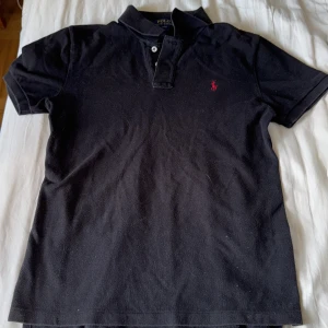 Svart Polo Ralph Lauren t-shirt - Svart t-shirt från Polo Ralph Lauren med klassisk pikékrage och två knappar vid halsen. Röd broderad logga på bröstet. Tillverkad i mjuk bomull, perfekt för en chill och stilren look. Kortärmad och normal passform.