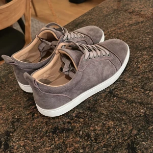 Grå mocka sneakers med vit sula - Snygga grå sneakers i mocka med vit platt sula och rund tå. Skorna har snörning i matchande grå färg och en röd detalj på undersidan av sulan. Insidan är ljusbeige och de har en stilren, minimalistisk design som passar till många olika outfits.