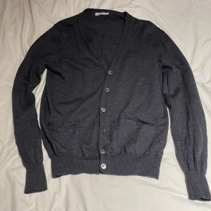 Mörkgrå kofta från Gran Sasso - Snygg mörkgrå kofta från Gran Sasso, tillverkad i Italien. Den har en klassisk v-ringning, knappar framtill och två fickor. Materialet är mjukt och stickat, perfekt för lager på lager. Passar dig som gillar stilrena och enkla plagg med en chill vibe.