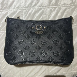 Svart axelväska från Guess med mönster - Snygg svart axelväska från Guess med präglat logomönster över hela väskan och guldfärgade detaljer. Väskan har dragkedja upptill och en extra ficka med dragkedja på baksidan. Perfekt storlek för det viktigaste och stilren design.
