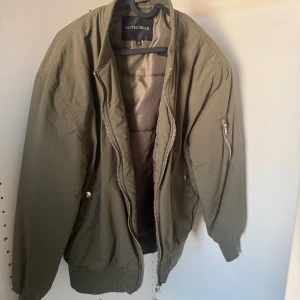 Olivgrön bomberjacka Rut&Circle - Snygg olivgrön bomberjacka från Rut&Circle i storlek 36. Jackan har klassisk ribbad krage, muddar och nederkant, samt dragkedja framtill och på ärmen. Perfekt för dig som gillar streetstyle och vill ha en chill look. Materialet är polyester och jackan är långärmad.