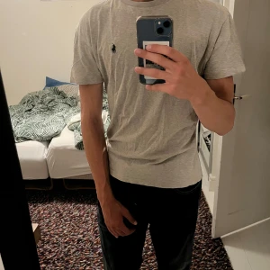 Grå t-shirt från Polo Ralph Lauren - Snygg grå t-shirt från Polo Ralph Lauren med klassisk rund hals och korta ärmar. T-shirten har en liten mörkblå Polo-logga broderad på bröstet. Mjuk och skön bomull som passar perfekt till jeans eller shorts. Enkel och stilren design.