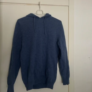 Merinoull hoodie - Blå merinoull hoodie med huva och snörning, perfekt för chill dagar. Tröjan har lång ärm och ribbade muddar vid ärmslut och nederkant. Enkel och stilren design som funkar till jeans eller mjukisbyxor. Skön och mjuk känsla, passar dig som gillar avslappnad stil.