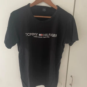 Svart t-shirt Tommy Hilfiger XL - Svart t-shirt från Tommy Hilfiger i storlek XL med klassisk rund hals och tryckt logga på bröstet. T-shirten är kortärmad och har en clean, stilren look. Materialet är mjuk bomull som känns skönt mot huden. Perfekt för dig som gillar ikoniska märken och enkel stil. Säljs för att den inte kommer till användning då den är för stor på än själv så tycker nån annan borde kunna utnyttja mer än mig. ABS! Pris går att diskutera.