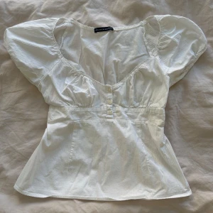 Vit babydoll top från Brandy Melville - Supersöt vit blus från Brandy Melville med puffiga korta ärmar och v-ringad hals. Blusen har knappar framtill och lätt rynkade detaljer vid axlar och byst. Tillverkad i mjuk bomull, perfekt för en chill och feminin look. OBS! Liten fläck längst ner men inget man ser om man har på sig den (se bild 3)