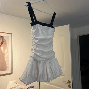 Vit kortklänning med svarta axelband - Supersöt vit kortklänning från Zara med breda svarta axelband och rynkad struktur. Klänningen har en volangig kjol och är figurnära upptill. Perfekt för dig som gillar en trendig och lekfull stil. Materialet känns lätt och har en snygg textur. Helt ny 