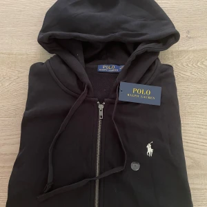 Svart hoodie från Polo Ralph Lauren -  Helt ny (alldrig andvänt) ingår scan, svart hoodie från Polo Ralph Lauren med klassisk broderad logga på bröstet. Tröjan har dragkedja framtill, huva med snörning och två fickor. Materialet känns mjukt och skönt, perfekt för chill dagar. Snygg och stilren design som funkar till det mesta.