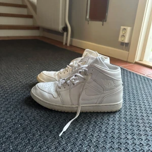 Nike Jordan 1 Mid Triple White - Säljer ett par Nike Jordan 1 Mid Triple White sneakers med klassisk high-top siluett. Skorna är helvita med perforerad tå, snörning och ikoniska Jordan Wings-loggan på sidan. Tillverkade i skinn för en clean och stilren look som alltid funkar.
