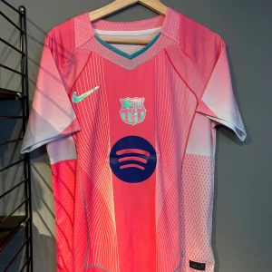 Barcelona Lamine Yamal #10 rosa tröja - Säljer en Barcelona fotbollströja med Lamine Yamal och nummer 10 på ryggen. Tröjan är från Nike och har en cool rosa färg med vita och ljusblå detaljer, samt tunna linjer som ger ett futuristiskt mönster. Materialet är lätt och andas, perfekt för match eller träning. Helt ny med tagg en replika 