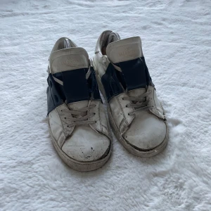 Valentino Open skor vit/blå - Säljer ett par Valentino Open skor i vit skinn med bred mörkblå rem över sidan. Klassisk rund tå, snörning och platt sula. Märket syns tydligt på tungan och sulan. Dem är väldigt slitna men har haft dem i två år och använt dem mycket. Sulan är väldigt sliten och den har lite slut märken men det går att få bort med att tvätta med sko tvål. Ny pris 7000kr