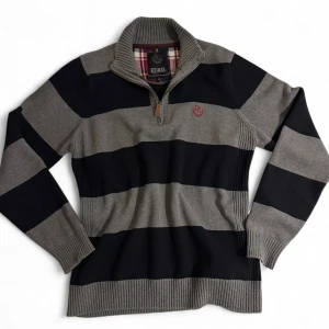 Randig half zip tröja från Etirel - Snygg stickad tröja från Etirel i XL med breda svarta och gråa ränder. Tröjan har en ribbad krage med half zip-dragkedja och ett broderat rött emblem på bröstet. Perfekt för dig som gillar sportig och klassisk stil.