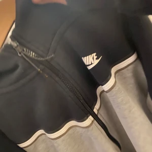 Nike tech huddiee - Nike hoodie med dragkedja i svart och grått, med vit logga på bröstet, har luva. Använd men i bra skick 