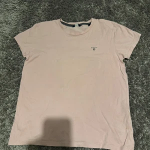 Ljusrosa t-shirt från GANT - Snygg ljusrosa t-shirt från GANT med diskret logga på bröstet. Klassisk rund hals och korta ärmar, perfekt för en chill och stilren look. Mjuk och skön bomull som känns nice mot huden. Enkel att matcha med jeans eller shorts. Storlek 158-164 men är samma som en XS