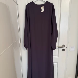 Mörklila abaya från El Bassira - Säljer en stilren långklänning från El Bassira i en mörklila ton. Klänningen har rund halsringning och långa ärmar med lätt puff vid handleden. Ny oanvänd abaya  Färg: mauve. Storlek 58