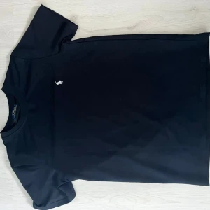 Svart t-shirt från Polo Ralph Lauren - Svart t-shirt från Polo Ralph Lauren med klassisk passform och rund hals. T-shirten har korta ärmar och en liten vit broderad logga på bröstet. Tillverkad i mjuk bomull som känns skön mot huden. Perfekt för en clean och stilren look.