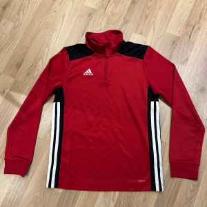 Röd Adidas långärmad tröja med half zip - Snygg röd Adidas tröja med svart och vita detaljer på axlar och sidor. Långärmad med half zip och hög krage, perfekt för träning eller chill. Liten reva på bröstet som syns lite 