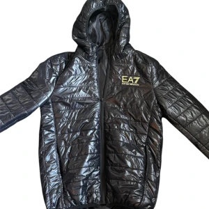 Svart pufferjacka EA7 Emporio Armani - Svart pufferjacka från EA7 Emporio Armani med glansig finish och quiltad design. Jackan har huva, hel dragkedja och EA7-logga i gult på bröstet. Perfekt för dig som gillar sportig stil och vill ha något som sticker ut i mängden.