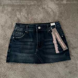 Mörk minijeanskjol från Missguided - Aldrig använd. Snygg mörkblå jeanskjol från Missguided med klassisk femficksdesign, bälteshällor och rå kant nedtill. Kjolen har en slim passform och är perfekt för dig som gillar en enkel men trendig look. Styla med din favorit-topp för en cool vibe.
