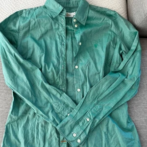 Grön skjorta från Morris Lady, strl 36 - Snygg grön skjorta från Morris Lady i storlek 36. Skjortan har klassisk krage, vita knappar och lång ärm. Materialet är bomull och på bröstet finns en broderad detalj med Morris-loggan. Perfekt för dig som gillar stilrena och färgglada plagg. Skjortan har en liten ”fläck” vid framsidan av knapparna, som jag inte lyckas få bort men den är inte så synlig. 