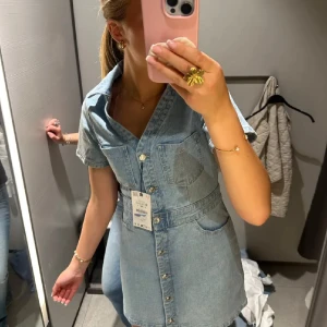 Kort jeansklänning med knappar - Snygg ljusblå jeansklänning med kort ärm, v-ringning och knappar hela vägen framtill. Toppen skick! Från Zara, i xs