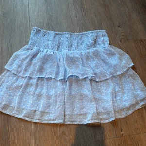 Blå volangkjol från Vero Moda - Supersöt ljusblå kortkjol från Vero Moda med volanger och smockad midja. Kjolen har ett diskret leopardmönster i vitt och är gjord i ett lätt och luftigt syntetmaterial. Perfekt för sommarens alla äventyr!