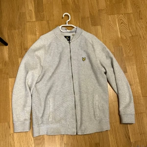 Grå kofta från Lyle & Scott - Säljer en stilren grå kofta från Lyle & Scott med dragkedja framtill och deras klassiska gula fågel-logga på bröstet. Tröjan har långärmade raglanärmar och två fickor. Perfekt för chill dagar eller när du vill ha en clean look.