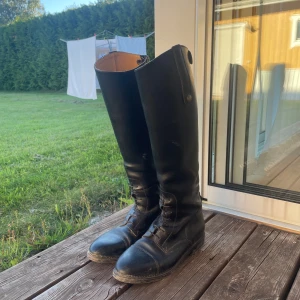 Svarta ridstövlar med snörning - Säljer ett par svarta ridstövlar med snörning framtill och dragkedja baktill. Stövlarna har rund tå och platt sula, samt knappdetaljer upptill. Perfekta för dig som gillar klassisk ridstil och vill ha ett par höga stövlar med snygg finish. SKA PUTSAS!!!