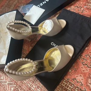 Champagnefärgade Chanel pumps med pärlor - Lyxiga pumps från Chanel i champagnefärgad satin med öppen tå och elegant klack. Skorna har en unik pärlbeströdd rem runt ankeln och guldfärgad innersula. Perfekta för dig som vill sticka ut med exklusiv design och detaljer.