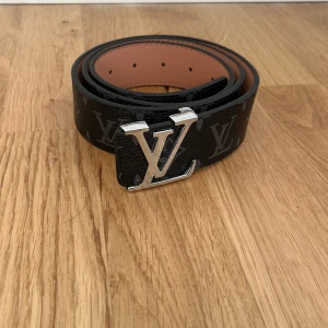 Louis Vuitton svart skärp med LV-spänne - Snyggt svart bälte från Louis Vuitton med klassiskt LV-monogram och silverfärgat LV-spänne. Bältet är gjort i exklusivt skinn och har en stilren design med bruna insidor. Perfekt accessoar för att lyfta din outfit med en lyxig touch.