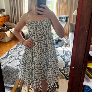 Vit blommig sommarklänning med tunna band - Supersöt vit kortklänning med blått blommönster och tunna axelband. Klänningen har en luftig passform, volangdetaljer och öppen rygg. Den har även en underkjol som gör den mindre genomskinlig💗