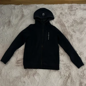Svart Sail Racing hoodiejacka strl 160 - Svart hoodiejacka från Sail Racing med dragkedja och huva. Den är lite blekt på vissa ställen (kolla bild 3 och 4) men annars bra skick.