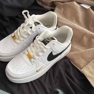 Nike Air Force 1 vit/svart med gulddetaljer - Snygga Nike Air Force 1 sneakers i vitt skinn med svart swoosh och gulddetaljer på snörningen och loggan. Klassisk låg modell med perforerad tå och vit platt sula. Perfekta för dig som vill ha en clean och tidlös look med lite extra bling. Säljer de för att de var för små byter gärna om ni har bra skor med bra skick i storlek 37,5 eller 38