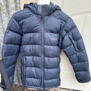 Mörkblå Peak Performance dunjacka - Snygg mörkblå dunjacka från Peak Performance med huva och dragkedja framtill. Jackan har en clean look med diskret logga på bröstet. Perfekt för kalla dagar och enkel att matcha med det mesta. Storlek 170!