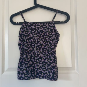Blommig linnetopp från Lindex - Supersöt linnetopp från Lindex med smala axelband och svart botten täckt av små lila och gula blommor. Perfekt för dig som gillar en feminin och lekfull stil. Tunt och mjukt material som känns skönt mot huden.