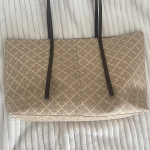 Beige shoppingväska från By Malene Birger - Snygg shoppingväska från By Malene Birger i beige med vitt rutmönster och långa mörkbruna axelband. Väskan är rymlig och har en innerficka med samma mönster. Tillverkad i tåligt syntetmaterial med stilren design.